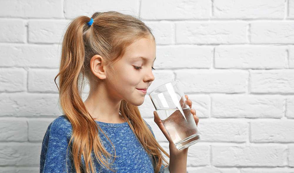 ילדה עם קוקיות עומדת ליד קיר ומביטה לתוך כוס מים