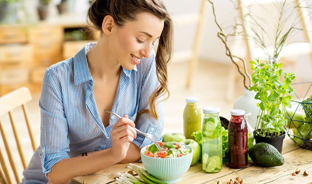 בחורה יושבת ליד שולחן ואוכלת סלט ירקות, ליד הקערה מונחים רטבים שונים