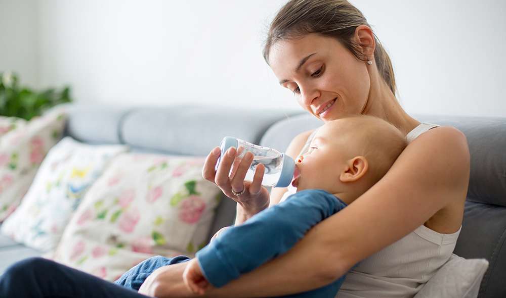 אם צעירה יושבת על ספה ונותנת לבנה התינוק לשתות מים מבקבוק